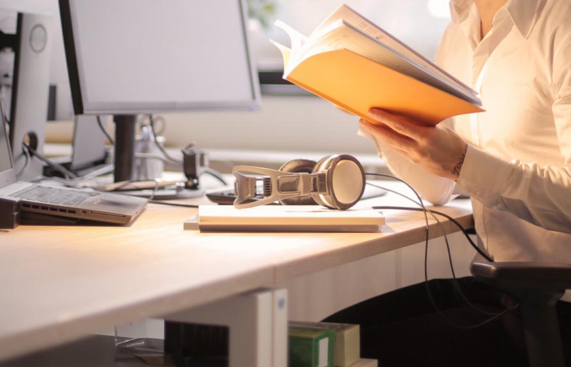 Closeup of a female holding a book while sitting at a computer station with headphones on the desk.