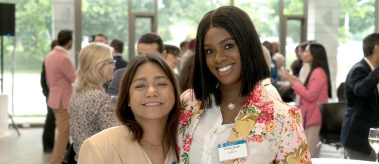 Two women posing and smiling for the camera, with others in the background mingling. 