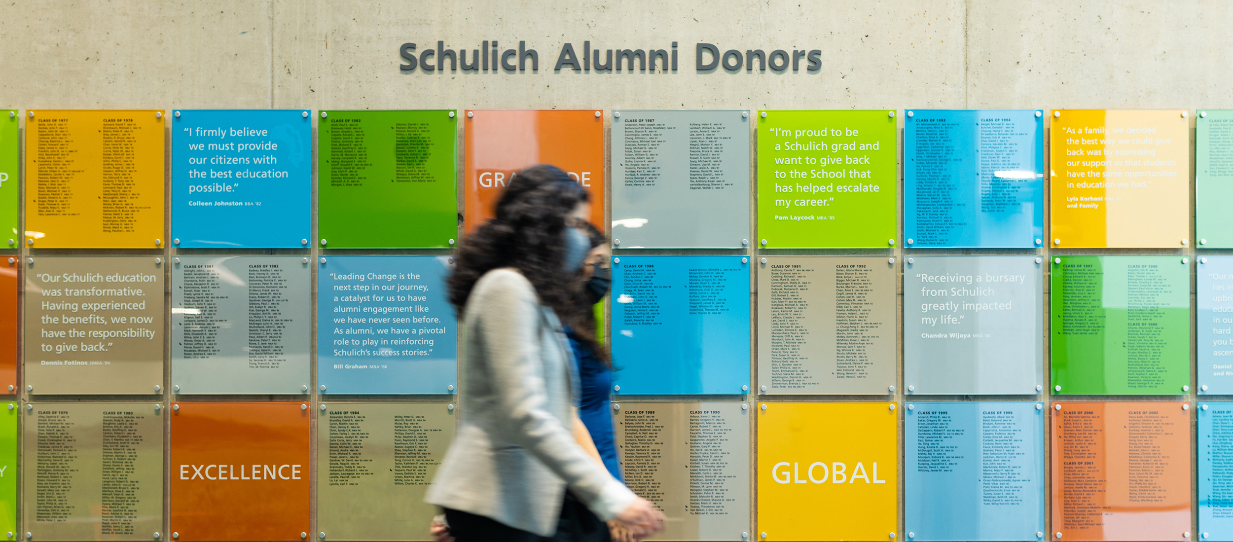 Two people walking by the multi-coloured Schulich Alumni Donor wall.