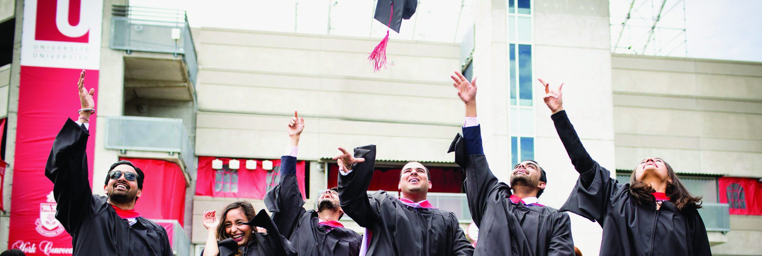 Graduates wearing gowns throwing mortarboard caps in the air, with York University signage in the background.