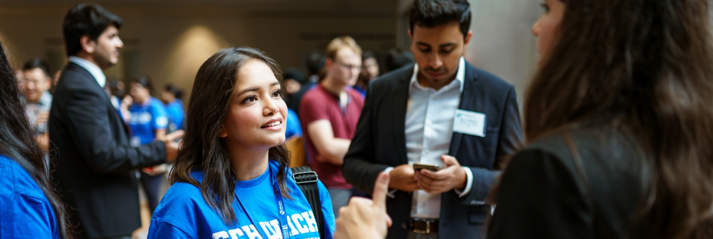 Focus on a female wearing a blue “Schulich” t-shirt, in conversation with others.