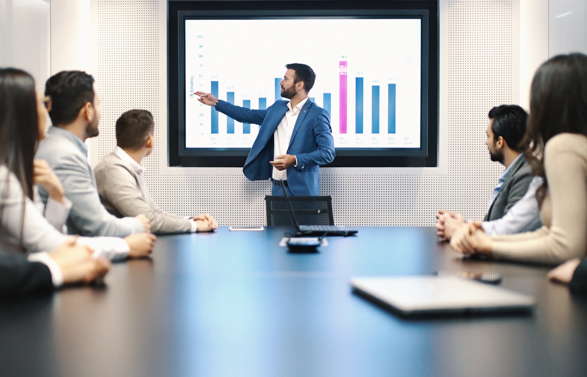 People gathered around a boardroom table, with one man standing in front of a video screen presenting a graph.