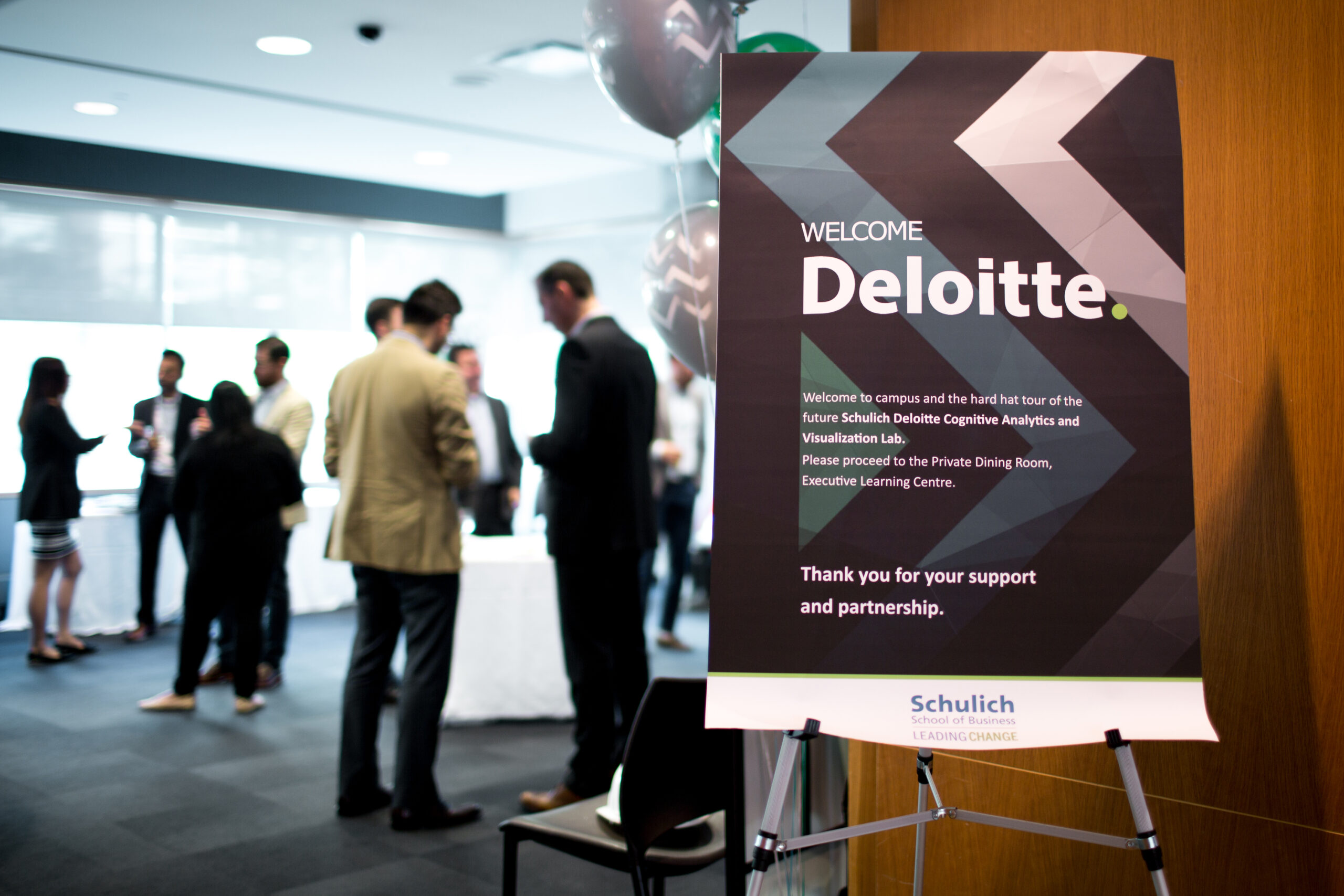 Closeup of a “Welcome Deloitte” sign with groups of people mingling in the background.